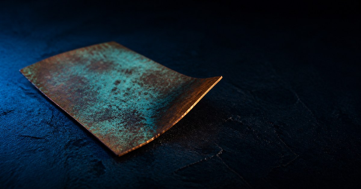 A single sheet of oxidized copper on dark stone, one lifted corner catching a cyan edge of light against warm patina